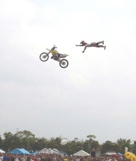 Una persona en competencias en moto en un sitio lleno de espectadores que miran cuando estando bien alto pierde la moto quedando como volando