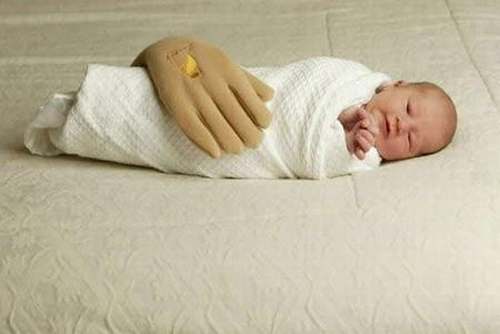 Vemos a un bebe que descansa en vuelto en una manta blanca pero luego tiene encima una almohada en forma de mano muy grande