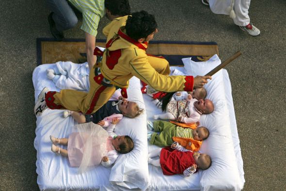 Tenemos a un hombre con un vestido amarillo con adornos rojos salta sobre unos bebes que stan el piso sobre unas cojines