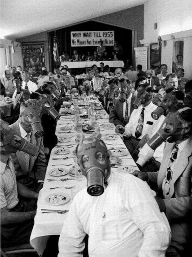 Vemos a una cantidad de personas en un salón muy grande en una mesa larga y de lado a lado  personas  con mascaras antigases   