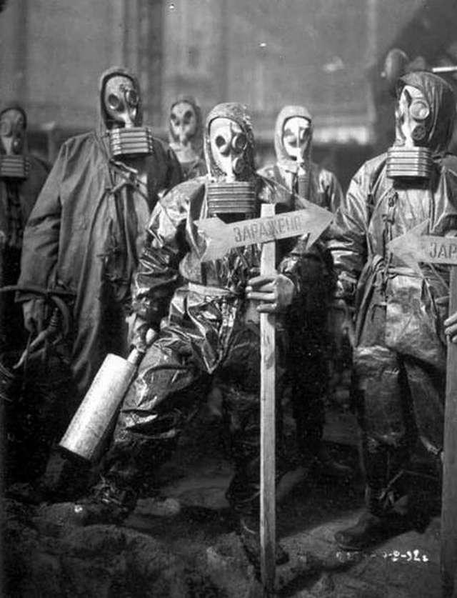Vemos seis hombres con abrigos y mascaras anti-gases antiguas uno tiene una flecha  que dice algo  