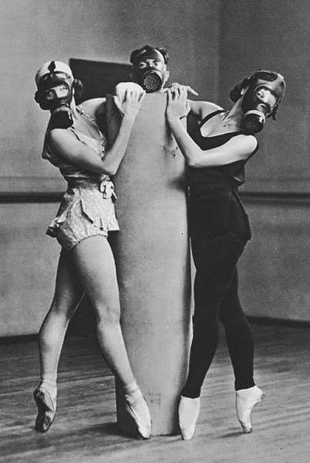 vemos a dos mujeres con  mascaras anti-gases   y un hombre en medio de ellas haciendo pasos de baile calzan zapatillas de Ballet  