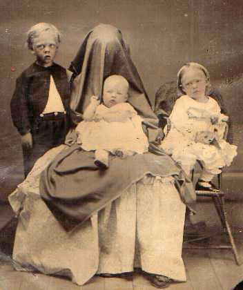 Tres hermosos niños dos niñas y un niño  vestidos de gala  miran fijamente algo  la niña grande esta sentada en una silla