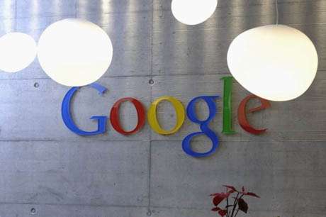 Vemos el logo de google  en colores sobre una parede gris y lamparas blancas