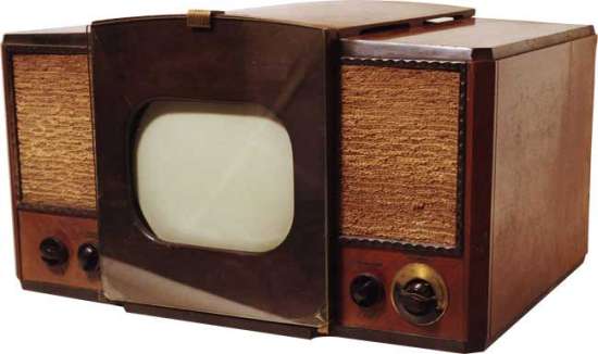 Vemos un televisor muy antiguo con pantalla muy pequeña dentro de un cajón de madera café oscura y las o tras partes en color habano colocado sobre una mesa