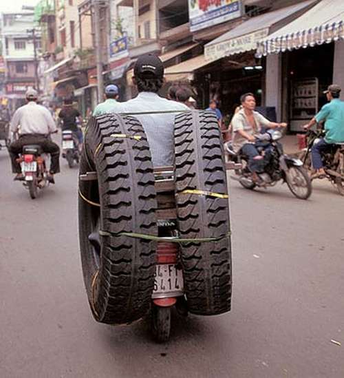 Un hombre en su moto transporta dos grandes llantas por una calle muy concurrida con mmas motos