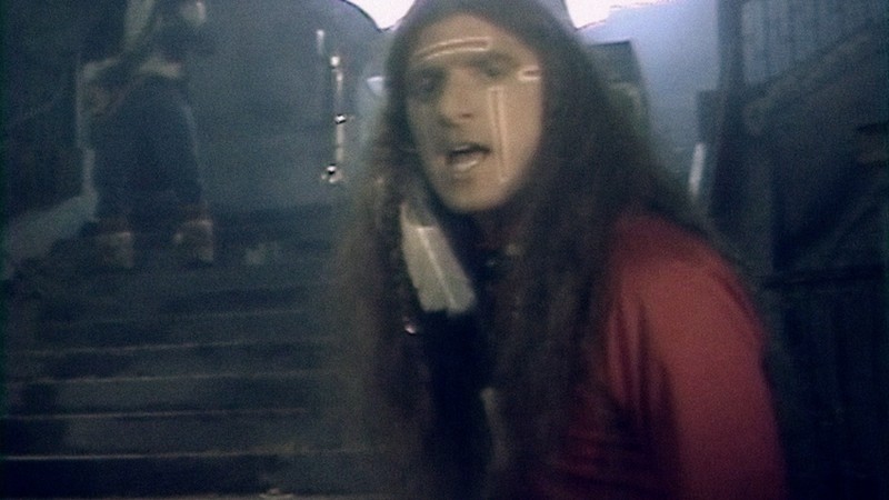 Una escena de un video musical en la que un músico de pelo largo mira a la pantalla con un bajista al fondo