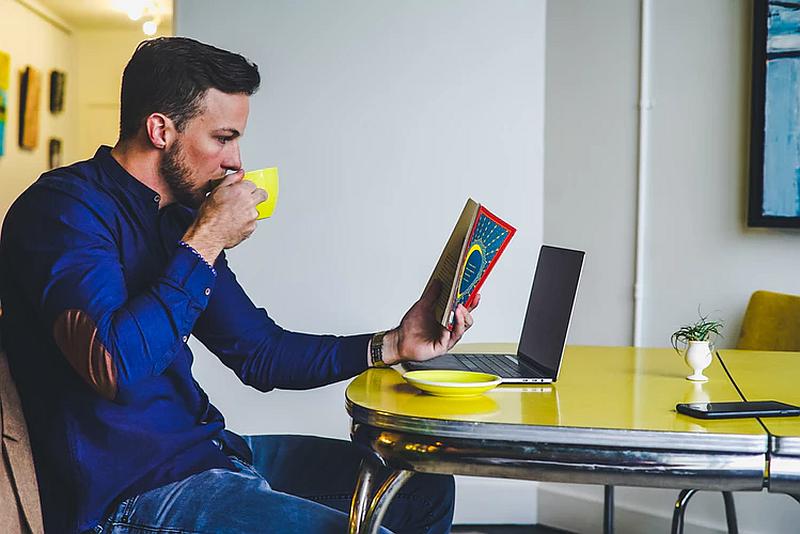 Vemos un hombre sentado frente a una mesa con un computador leyendo un libro y tomando un café