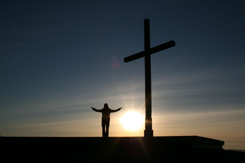 Vemos un lindo atardecer una cruz y una persona que levanta sus manos