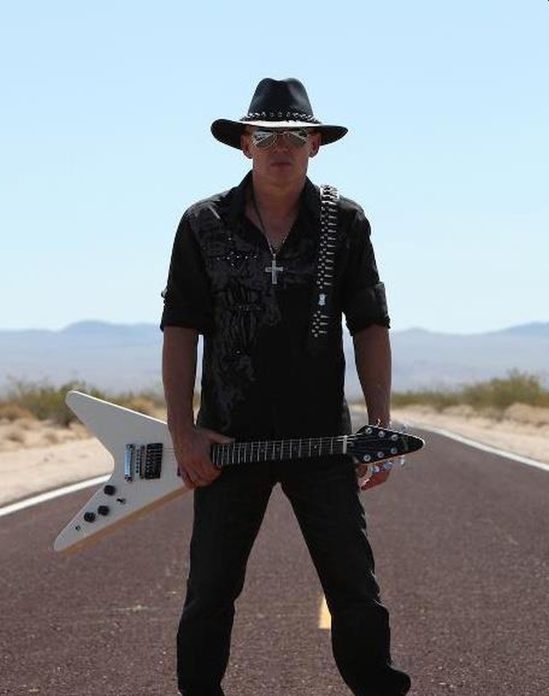 Un hombre vestido todo de negro con gafas y sombrero y una guitarra