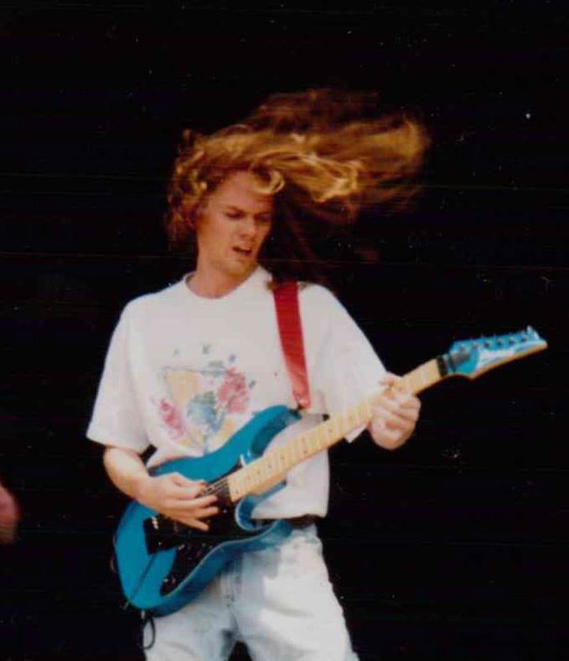 Un joven de pelo rubio y largo con ropa blanca interpreta su guitarra