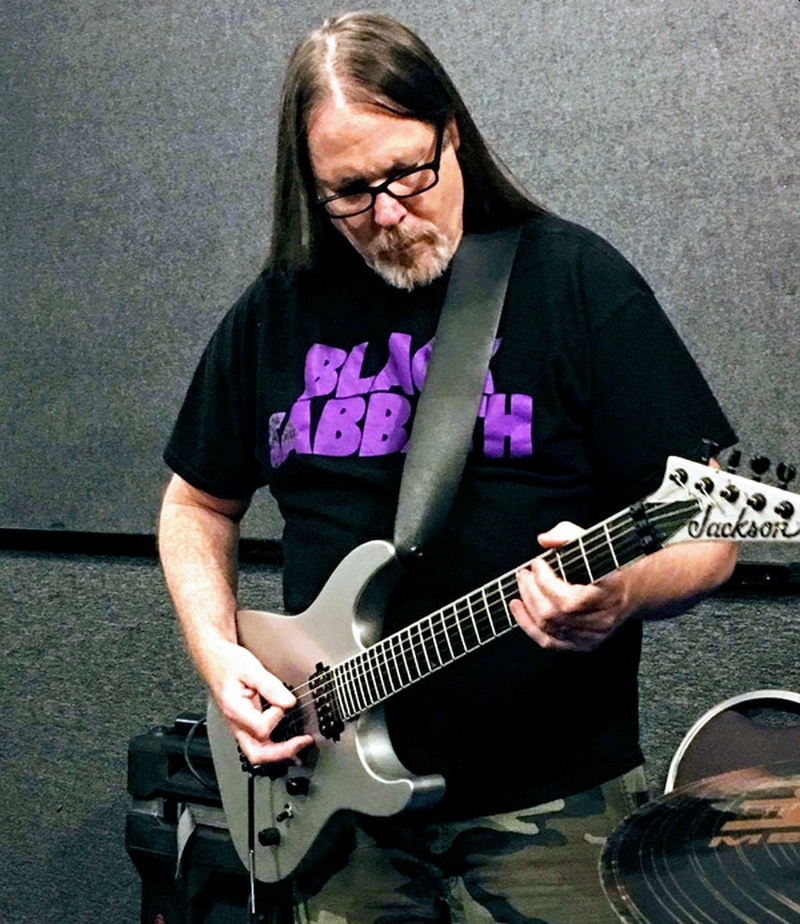 Un hombre mayor pelo largo y bigote  con lentes toca su guitarra