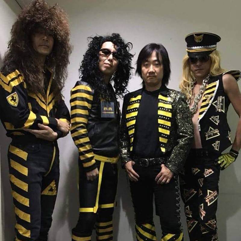 Cuatro integrantes de una banda con ropas negras con listas amarillas y triángulos y palabras