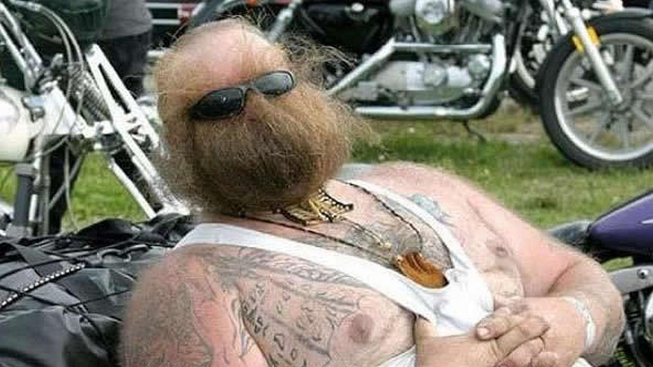 Un hombre con gafas y una barba que tapa toda su cara recostado en una moto