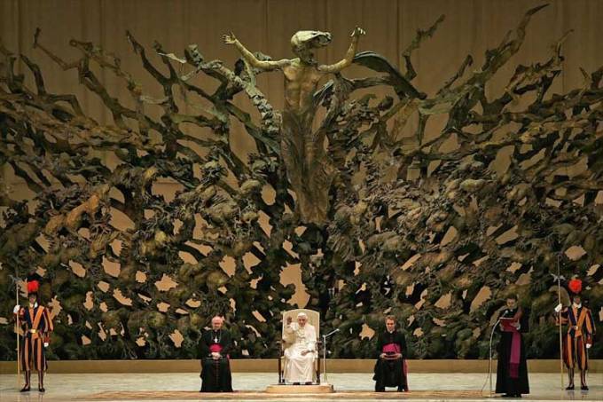 El Papa sentado y detrás una gran escultura conformada por calaberas brazoas pies, la escultura es larga y ancha y en la mitad de esta se encuentra jesus