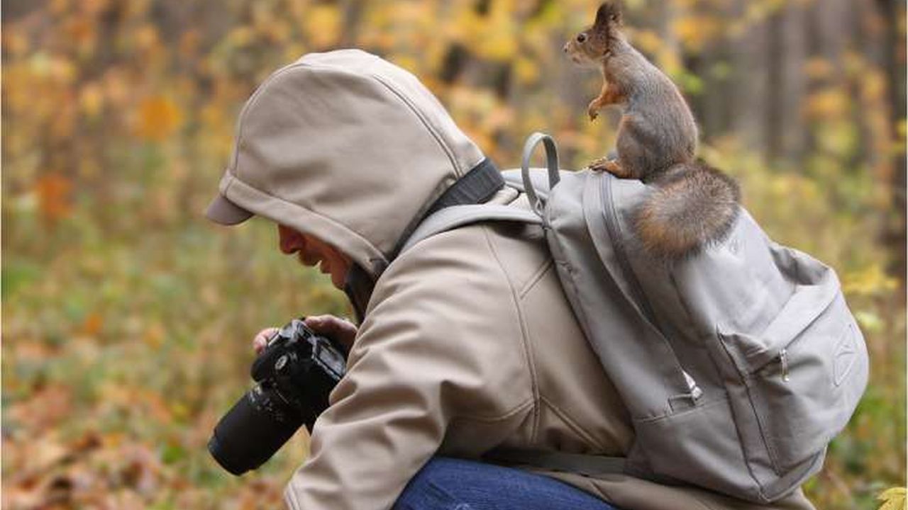 Un hombre tomando fotografías y encima de su mochila que la lleva en la espalda tiene montada una ardilla