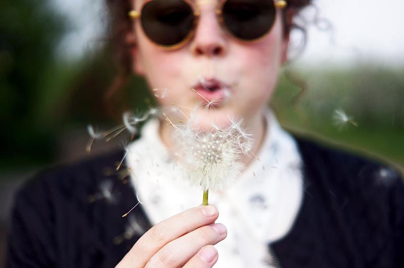 Un apersona joven con gafas deportivas sopla una flor silvestre  que derrama muchos pelillos