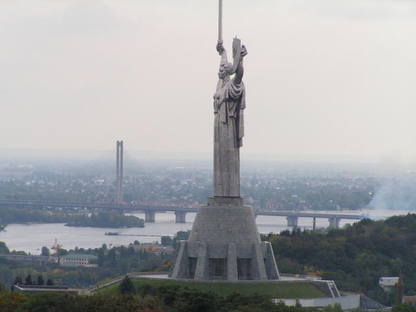 Figura femenina en color gris sobre un pedestal llevando una espada en su mano derecha se observa al fondo una gran ciudad
