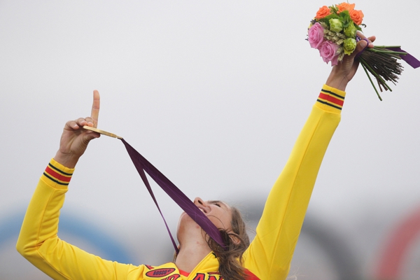 Una mujer joven con uniforme amarillo levanta su mano con una medalla que lleva en su cuello y tambien un ramo de flores