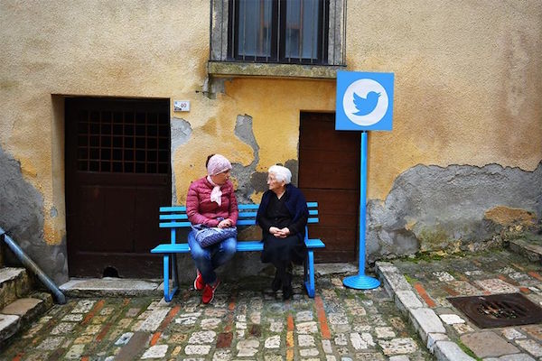 Dos personas sentadas en una banca azul y al lado un poste con el logo de twitter