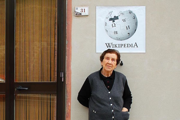 Una señora de edad recostada en la pared y sobre la cabeza de ella esta el logo de wikipedia