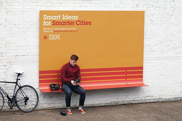 Un hombre sentado en una saliente que tiene un aviso gigante donde habla de la gente inteligente en ciudades inteligentes a su lado tiene una bici