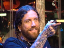 Un músico moreno con barba trenzas areta y tatuajes sonriente con un libro en sus mano