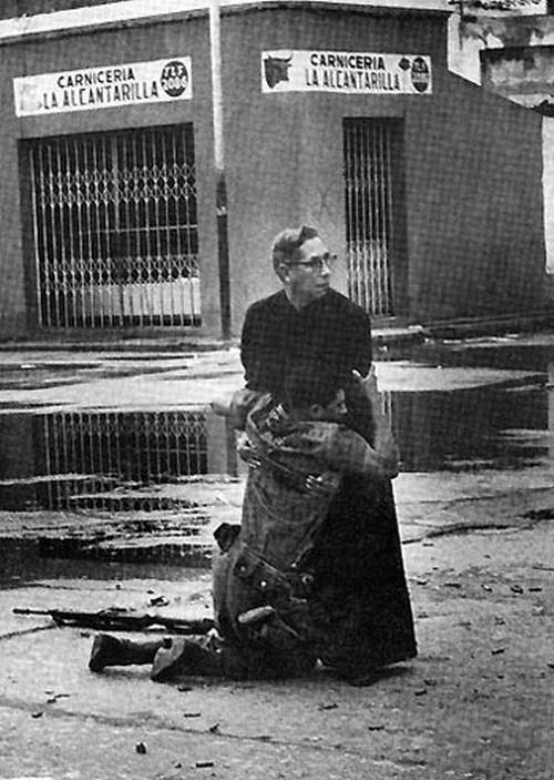 Tenemos un sacerdote al que se le abraza un soldado herido a los lados vemos edificios destruidos