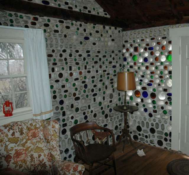 Vemos el interor de una casa hecha con botellas donde estan varias sillas mesas y una ventanaana y  en la pared se ven las botellas  