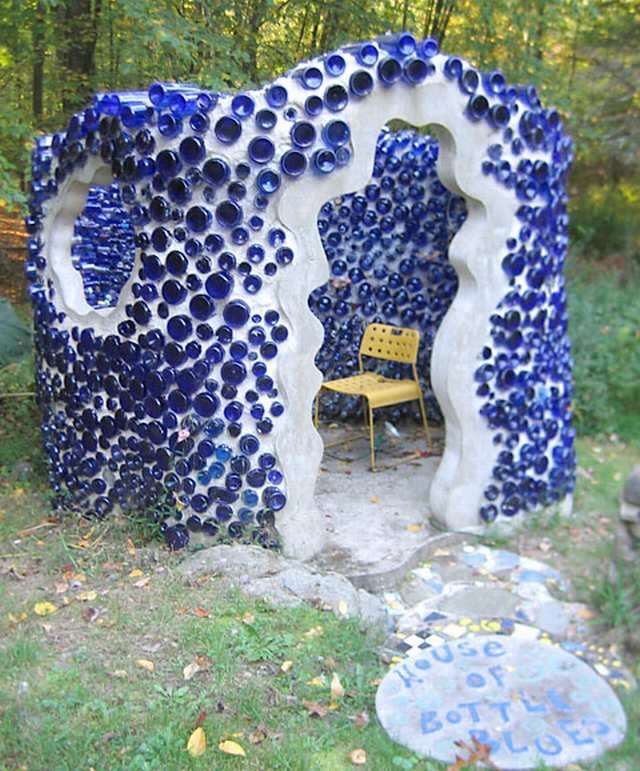 Vemos una estrutura blanca con botellas azules y dentro hay una silla ,amarilla  y en el piso sobre una piedra se lee la casa de las botellas azules 