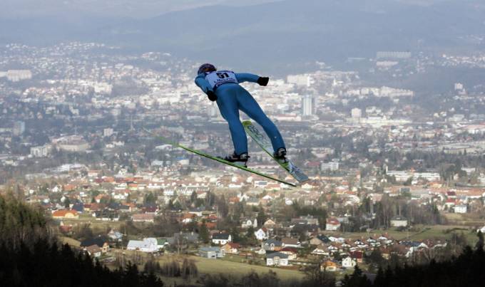 Un hombre con ski  hace un salto largo de donde se ve una ciudad al fondo