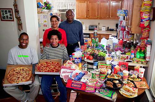 Vemos a una familia frente a mucha comida como pizzas paquetes leche soda queso mantequilla  frutas salsas  