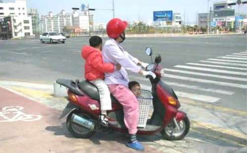 Una mujer conduce una moto con un niño atrás y un bebe en un canastillo plastico