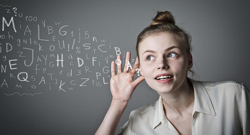 Vemos a una mujer rubia joven que colca su mano en el oído para  escuchar mejor frente a un tablero negro lleno de letras y numeros
