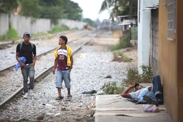 Ahora vemos a dos hombres muy jóvenes  que caminan al lado de la carrilera del tren y otro que descansa acostado en el piso