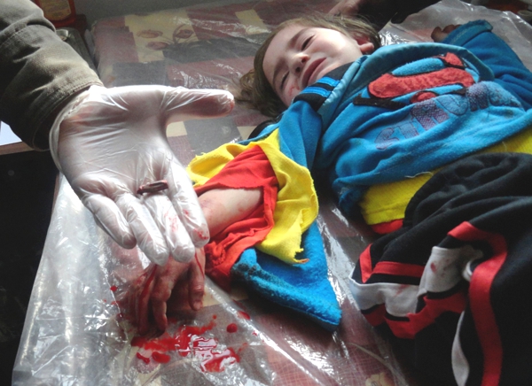 Una niña tendida en el suelo sobre un plastico y a su lado se ve una mano enguantada que muestra una bala la niña llora y tiene cara de dolor