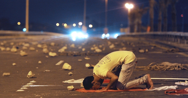Un hombre muy joven musulmán  ora inclinado sobre su alfombra y en el piso se ve la gran cantidad de piedra que hay despues de una protesta