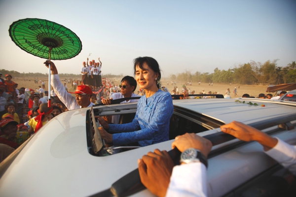 Una mujer en un lindo carro posa sonriente para una foto al lado un hombre lleva una sombrilla oriental en color verde a su lado hay una buena cantidad de gente 