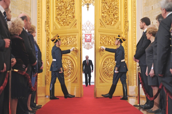 Vemos aun hombre de mediana estatura que entra a través de unas puertas doradas donde unos guardias uniformados lo reciben en la estancia se ven personas que lo esperan