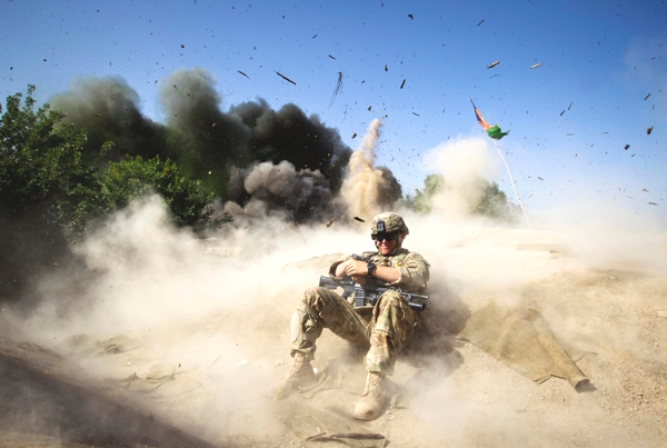 Un soldado con uniforme camuflado  esta sentado en el suelo de arena y atrás de e se ve una columna de arenal