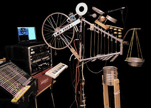 Un complejo instrumento donde se ve un aro de bicicleta utensilios de cocina un tarro de pintura campanas de teléfonos antiguos y teclados